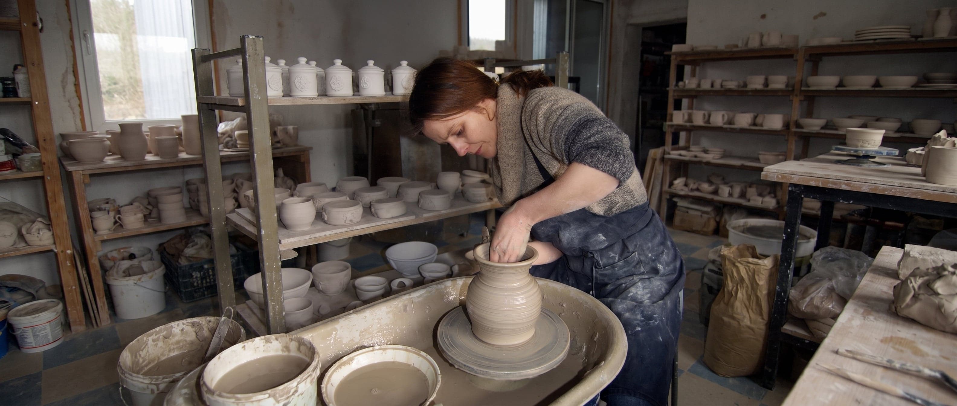 Brenda McGinn throwing a clay vase at the wheel in her Glaslough studio, shelves of drying pots around her.